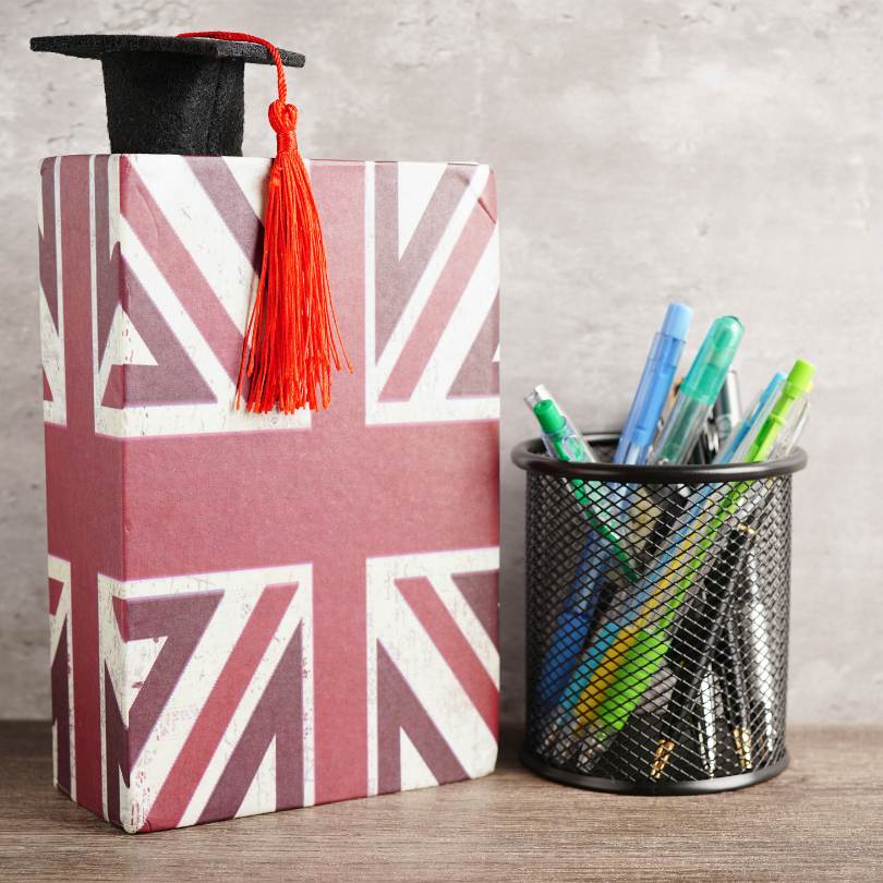 Libro cubierto con la bandera del Reino Unido y decorado con un birrete de graduación, junto a un portalápices con bolígrafos de colores