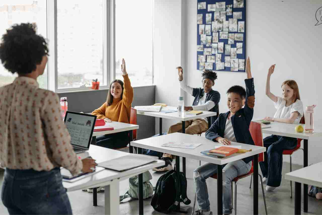 Estudiantes en un aula levantando la mano para participar, mientras una profesora les observa desde el frente, destacando un ambiente de aprendizaje activo y participativo