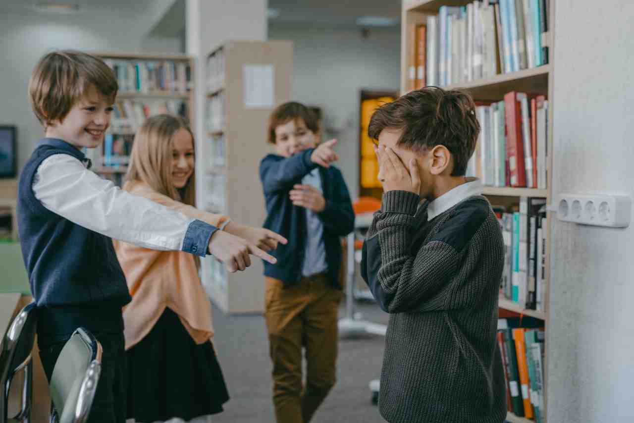 Grupo de niños en una biblioteca, riéndose y señalando mientras un niño cubre su rostro con las manos