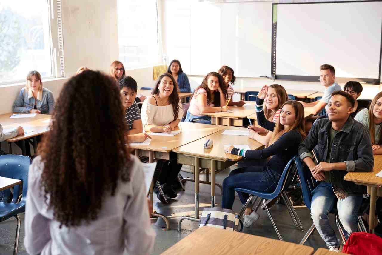 Grupo de estudiantes en un aula participando activamente mientras la profesora los guía desde el frente de la clase