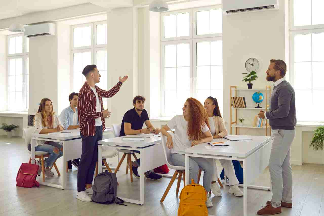 Estudiante participando activamente en clase mientras el profesor y otros compañeros lo escuchan en un aula luminosa