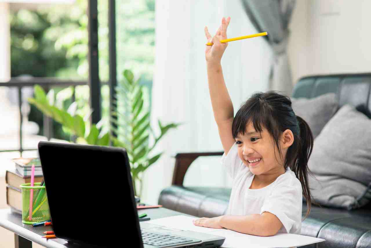 Niña levantando la mano con una sonrisa mientras participa en una clase virtual desde su laptop en casa