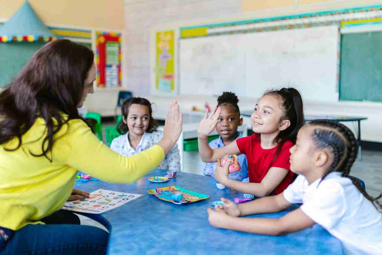 Maestra dando un 'choca esos cinco' a una niña en un aula de preescolar mientras otros niños observan, en un ambiente de aprendizaje lúdico