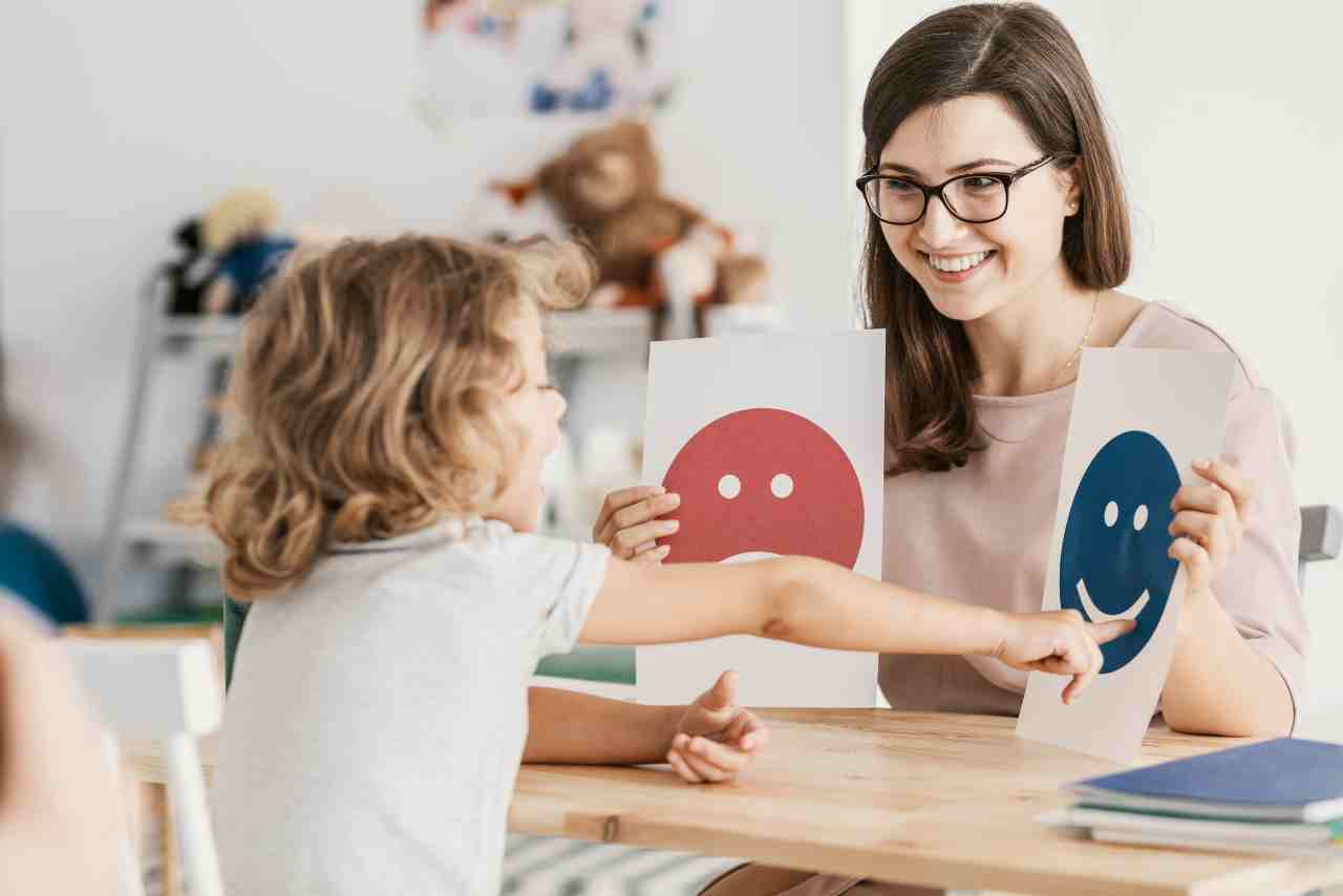 Niña eligiendo entre dos tarjetas con caras felices y tristes en una sesión educativa con una terapeuta, representando el aprendizaje emocional y el reconocimiento de emociones