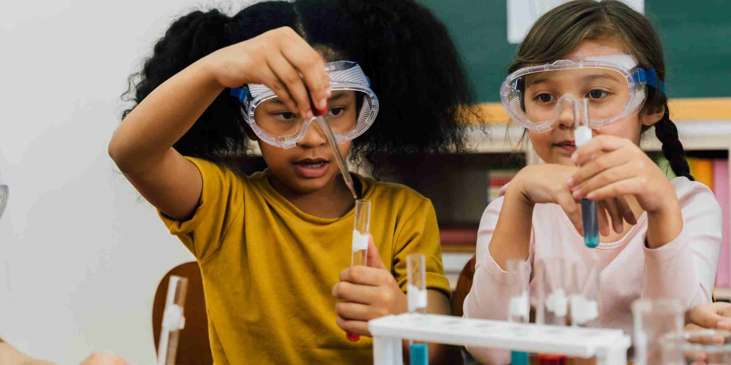 Niñas realizando un experimento de química en el aula, usando gafas de protección mientras manipulan tubos de ensayo