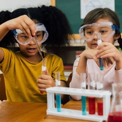 Niñas realizando un experimento de química en el aula, usando gafas de protección mientras manipulan tubos de ensayo con zoom