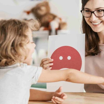 Terapeuta mostrando tarjetas de emociones, una con una cara triste y otra con una cara feliz, mientras un niño señala la tarjeta con la cara feliz